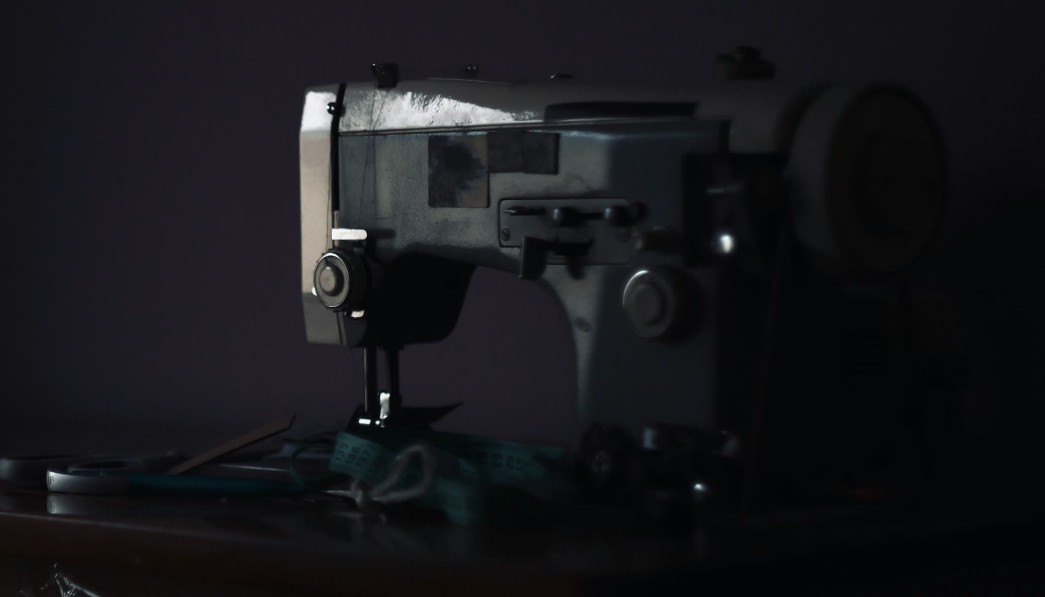 Máquina de costura desligada no escuro, com a fita métrica e a tesoura largadas na mesa do lado